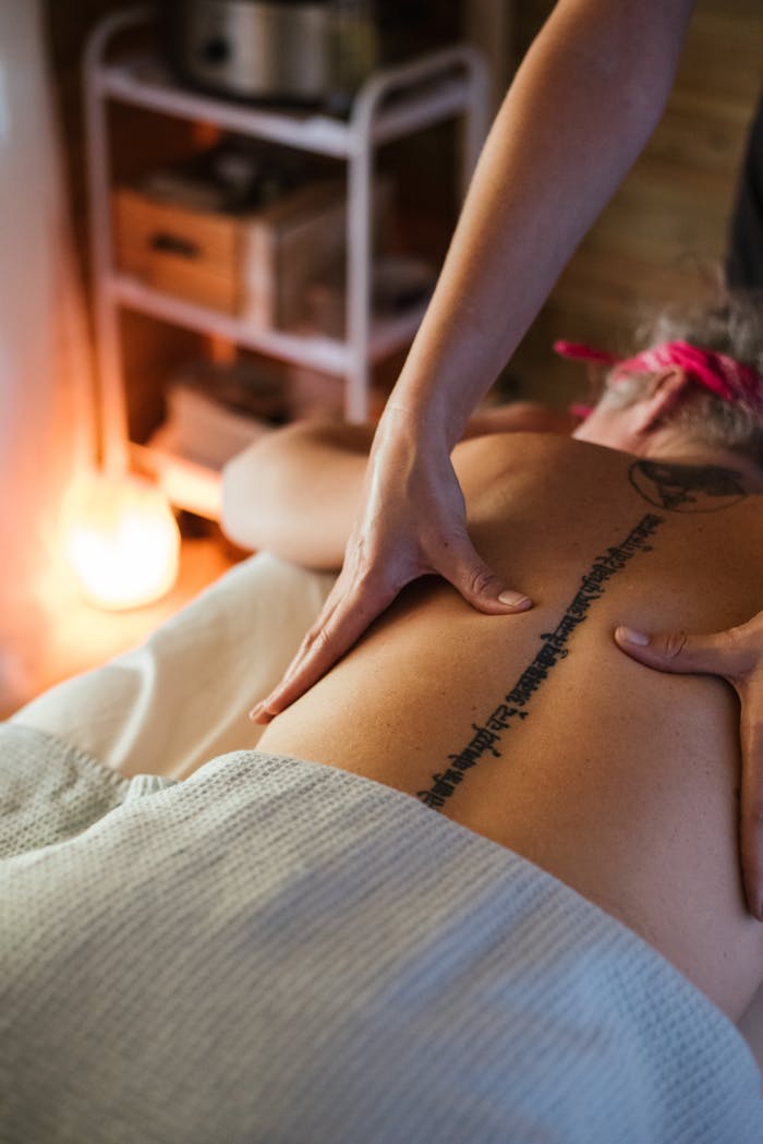 From above back view of unrecognizable tattooed female client with headband lying on stomach under blanket during massage session in spa on blurred background